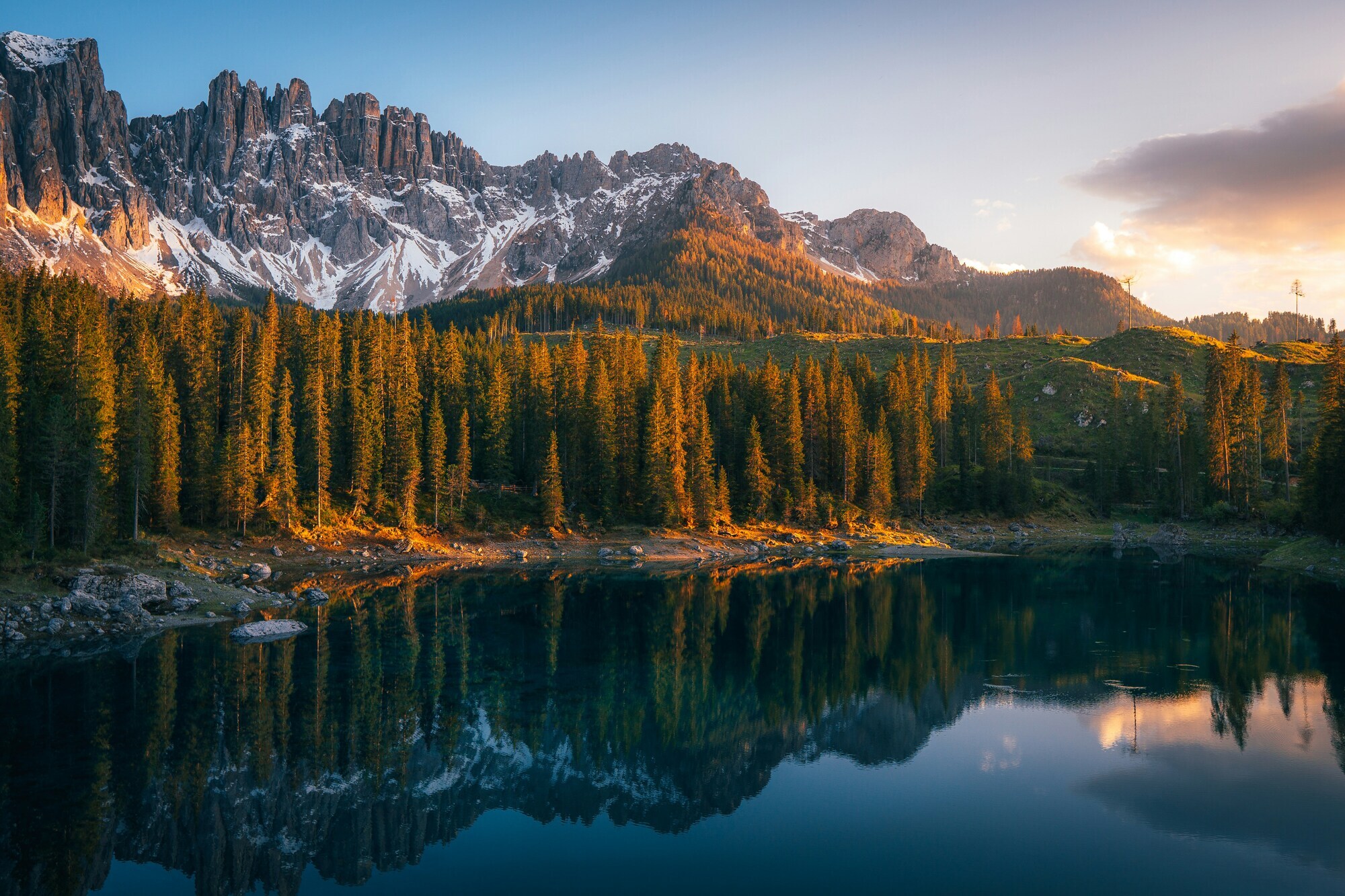 Evening scene at a dark lake, framed by trees in the sunset and a mountain range in the background