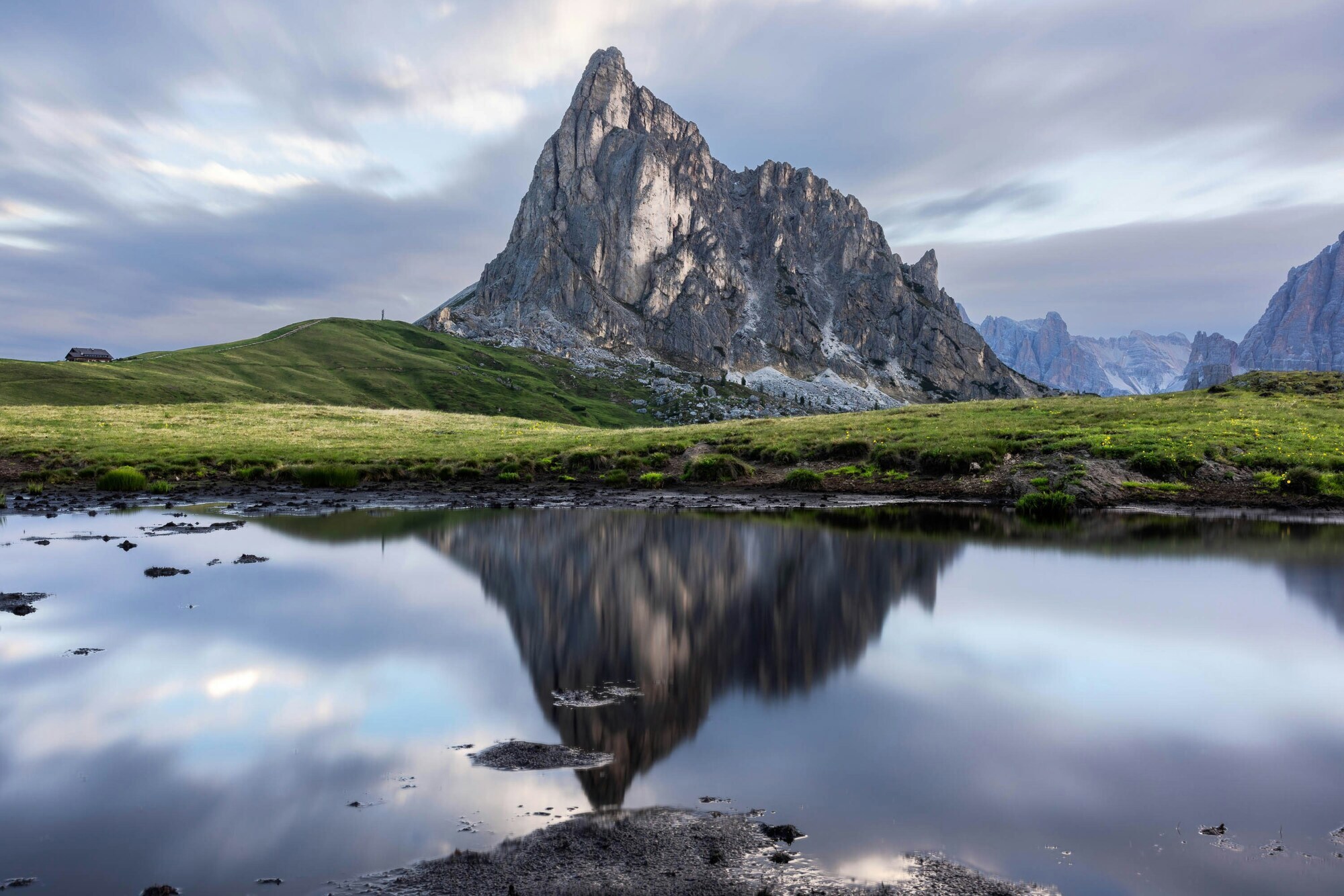 Mountain reflecting itself in a lake, framed by green meadows and cloudy sky