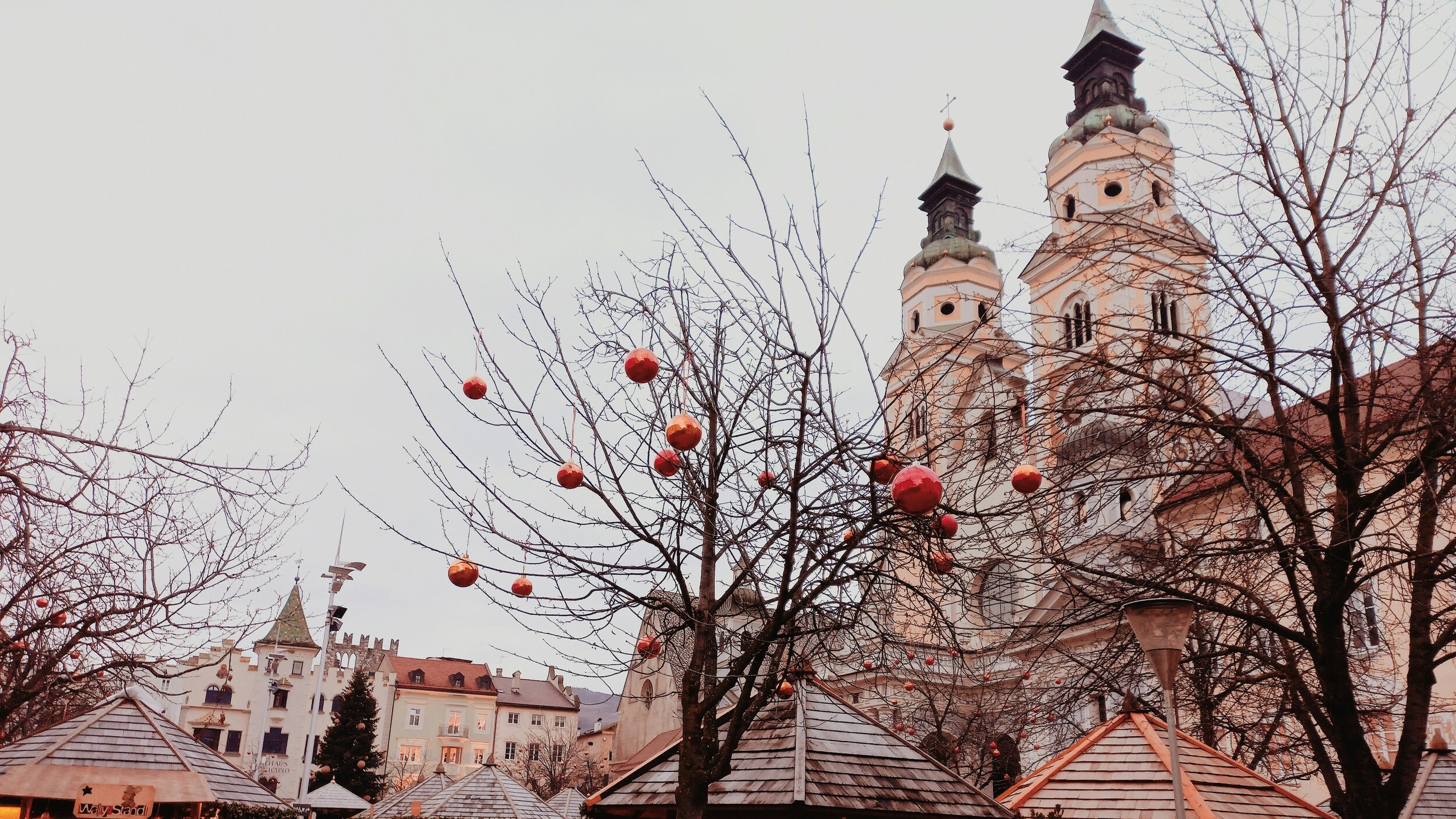 Bare tree decorated with red Christmas baubles between small wooden roofs and a city backdrop with church towers