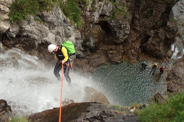 A man abseils over a waterfall, three people waiting below at the water basin