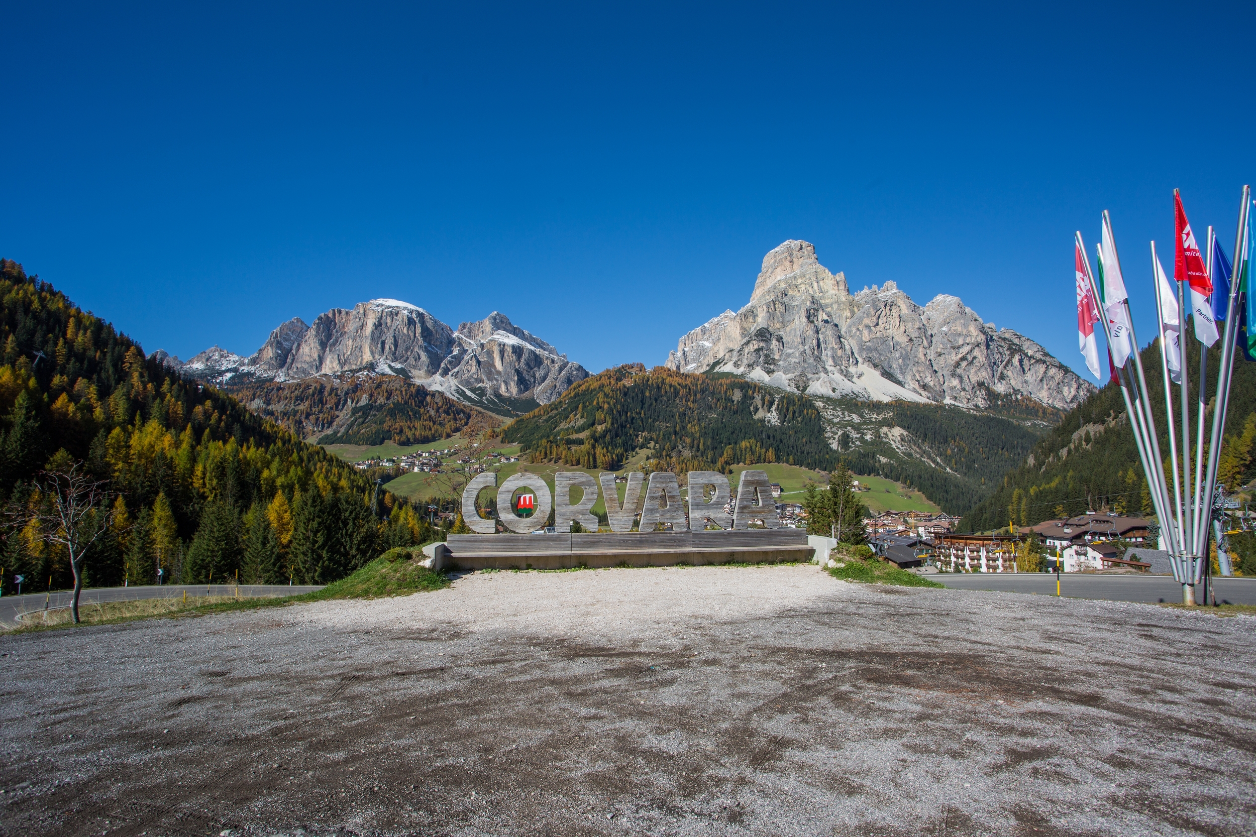 Wooden lettering "Corvara" in the middle of a mountain landscape, colourful flags on the right