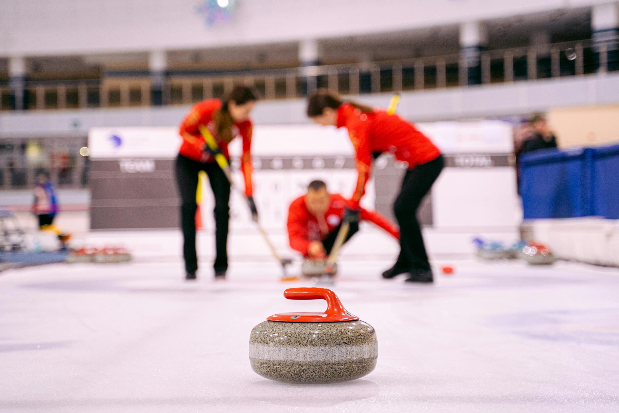 Curling stone with red handle, 3 players in red and black dress in blurred background