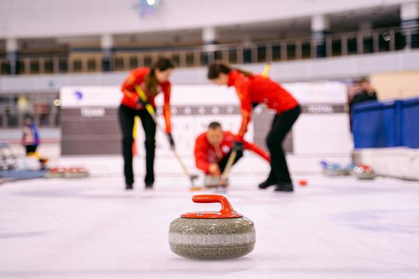 Curling stone with red handle, 3 players in red and black dress in blurred background