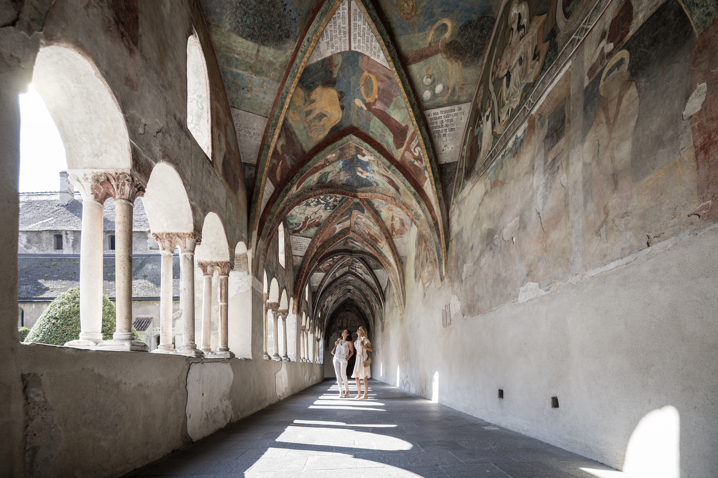 2 people in a cloister decorated with colourful frescoes, light coming in from outside