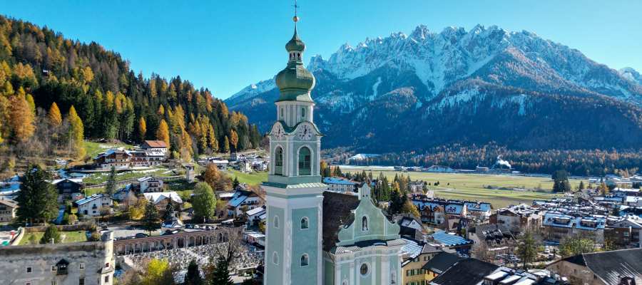 Light green church with tower, surrounded by houses, autumn-coloured trees and a snow-capped mountain