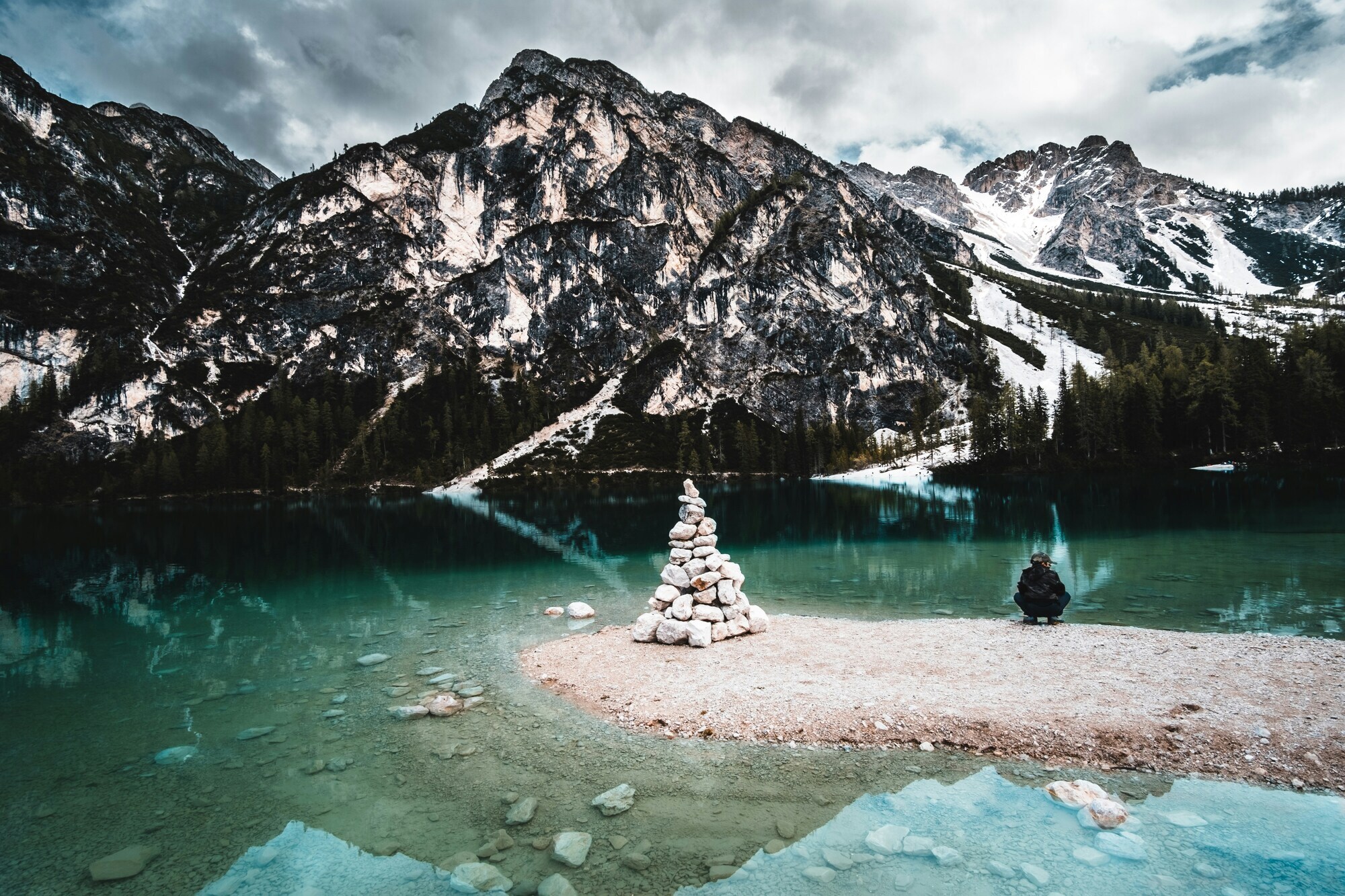 Child sitting next to a cairn on the shore of a crystal-clear lake, with snow-capped mountain peaks in the background