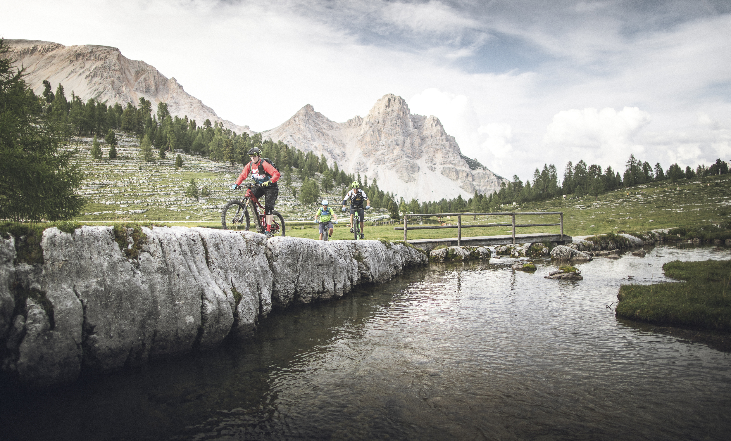 3 mountain bikers ride along a stream through a mountain landscape