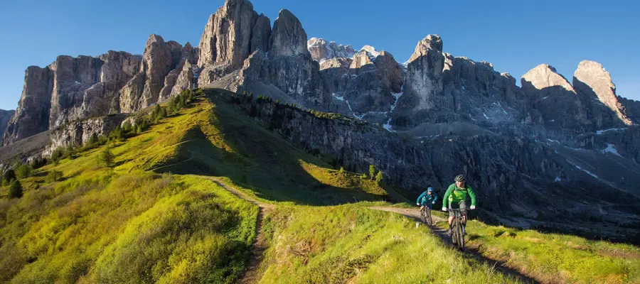 Bikers on a narrow path surrounded by meadows and mountains