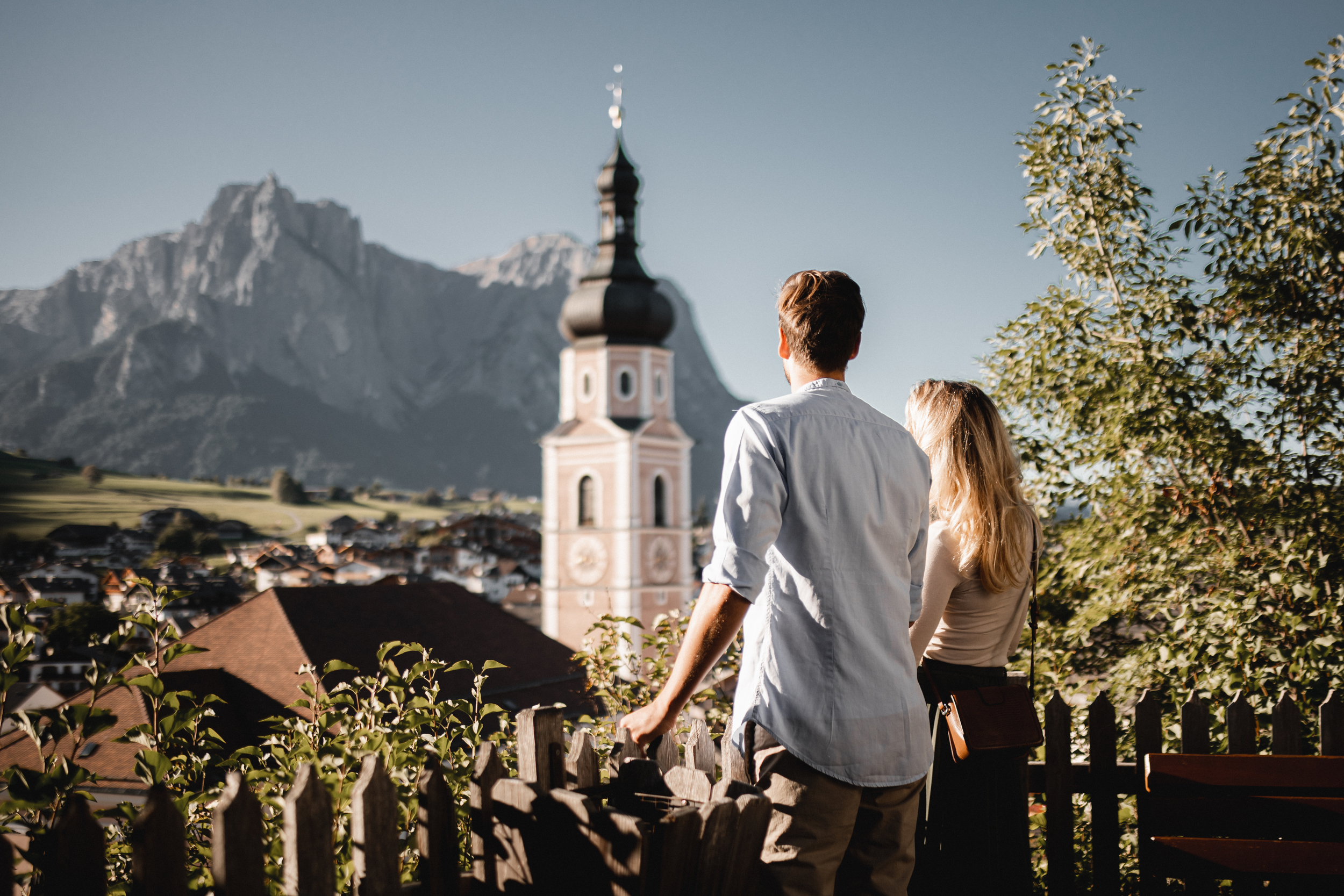 A couple looks away towards a village with a church tower in the centre and a mountain range in the background